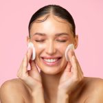 Home daily skincare. excited young lady cleaning face with cotton pads and smiling, doing skin care routine over pink background